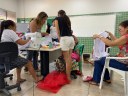 Para cego ver: mulheres distribuídas em uma sala, costuram roupas. 
